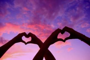 A couple's hands make heart shapes while held up to a beautiful sunset sky